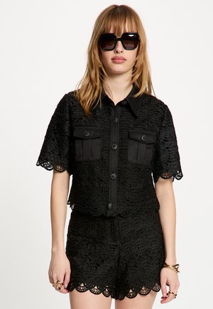Mujer con camisa negra de encaje de manga corta abotonada con dos bolsillos frontales y shorts a juego de encaje, complementados con gafas de sol negras y joyas de oro.