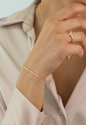 Gold bracelet with linked clear stones, featuring a delicate chain. Worn on a hand with matching rings, against a light satin blouse.