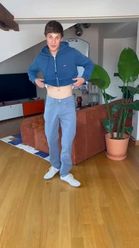 Sudadera corta azul, pantalones azul claro y zapatillas blancas. Suelo de madera natural y plantas verdes en el fondo.