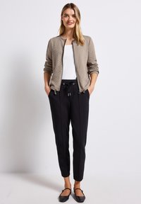 Vadderad beige bomberjacka med dragkedja, rund halsringning och elastiska ärmslut, i kombination med smalrandiga svarta byxor och platta skor.