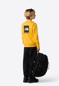 Gul sweatshirt med svart grafisk logo på baksiden, svarte bukser, og holder en svart ryggsekk. Skoene er svarte med teksturerte såler.