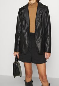 Schwarze Blazer aus Kunstleder mit Reverskragen, zwei_frontlichen Taschen und einem gefütterten Innenfutter, kombiniert mit einem braunen gerippten Top und schwarzen Shorts.