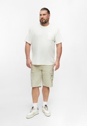 Man met baard die een wit T-shirt met korte mouwen draagt, beige cargoshorts, witte sokken en witte sneakers, staand tegen een effen achtergrond.