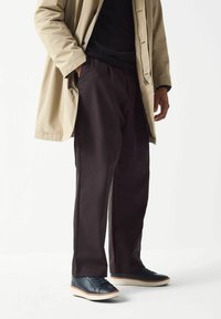 Cappotto impermeabile beige sopra un maglione nero, abbinato a pantaloni marroni scuri e sneakers blu con suole bianche e dettagli marroni.