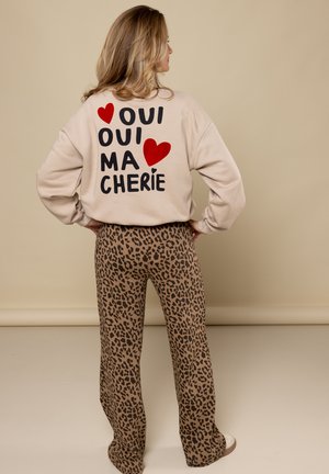 Beige sweatshirt met zwarte en rode tekst, "OUI OUI MA CHÉRIE," gecombineerd met bruine panterprintbroek en witte sneakers.