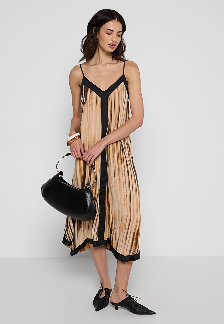Beżowa sukienka w kształcie litery A na ramiączkach typu spaghetti z czarnymi pionowymi paskami. Czarny detal przy dekolcie i u podstawy. Modelka trzyma zakrzywioną czarną torebkę.