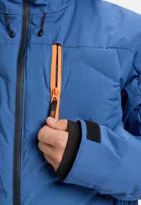 Hand, die den orangefarbenen Reißverschluss an einer blauen Outdoor-Jacke mit schwarzen Innenbündchen und einem Klettverschluss am Ärmel anpasst.