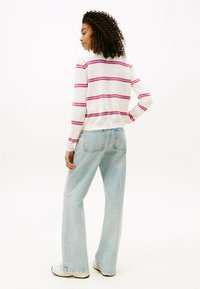 Jeune femme avec des cheveux bouclés portant un pull blanc à rayures rouges, un jean large bleu clair et des baskets blanches, se tenant de profil.