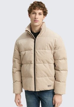 Joven con chaqueta acolchada beige de cuello alto y cremallera frontal, combinada con jeans azules, de pie contra un fondo liso.