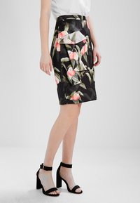 Žena v černé sukně po kolena s růžovým, broskvovým a zeleným vzorem listů, s volánkovým detailem, bílé blůze a černých sandálech na vysokém podpatku.