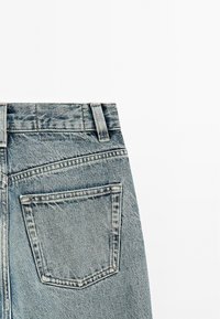 Jeans in denim blu sbiadito con vita alta, cinque passanti per cintura, una tasca sul retro e dettagli di texture visibili sul tessuto.