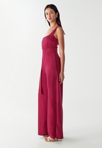 Vrouw staat in een mouwloze, donkerroze jumpsuit met brede pijpen, een vierkante halslijn en een strikceintuur, met beige sleehakken aan.