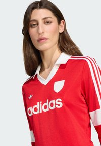 Rød fotballtrøye med hvite detaljer, med v-hals, et emblem på brystet og tre hvite striper nedover ermene.