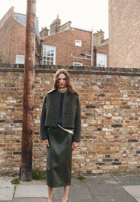 Groene wollen-blend cropped jacket met een textuurpatroon, gecombineerd met een diep groene leren midi-rok. Bakstenen muur op de achtergrond.