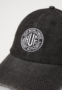 Čierna denimbová čiapka s oválnym šiltom, obsahujúca okrúhly biely vyšívaný logo s nápisom "HUF WORLDWIDE TRANSIT AUTHORITY."
