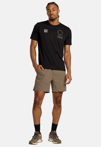 T-shirt nera a maniche corte con logo sul lato sinistro del petto, abbinata a pantaloni beigi. Il modello indossa calze scure e scarpe da trekking marroni con accenti colorati.