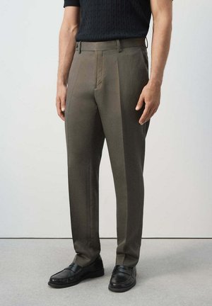 Uomo che indossa pantaloni sartoriali verde oliva e mocassini neri, in piedi davanti a uno sfondo chiaro e uniforme.