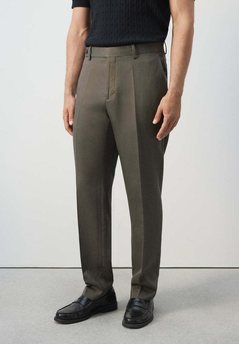 Homme portant un pantalon sur mesure vert olive et des mocassins noirs, debout devant un fond clair uni.