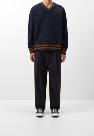 Navy v-neck sweater with contrasting orange striped ribbed hem, paired with dark trousers and black sneakers. Smooth knit texture.