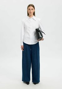 Camicia bianca con colletto a punta, abbinata a jeans in denim blu a gamba larga. Il modello tiene una borsa nera strutturata.
