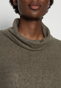 Olivgrünes, weiches Stricktop mit einem Schalkragen und strukturiertem Stoff, der ein dezentes Muster auf dem Oberteil aufweist. Klare, schlichte Design-Details.