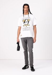 Una maglietta grafica bianca presenta il logo degli "Anaheim Ducks", abbinata a jeans grigi slim-fit. Il modello tiene in mano una piccola borsa nera e indossa scarpe da ginnastica nere.