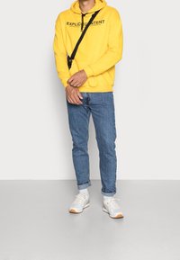 Żółta bluza z kapturem z czarnym nadrukowanym tekstem, połączona z jasnoniebieskimi dżinsami podwiniętymi na mankietach oraz białymi trampkami z pomarańczowymi akcentami.