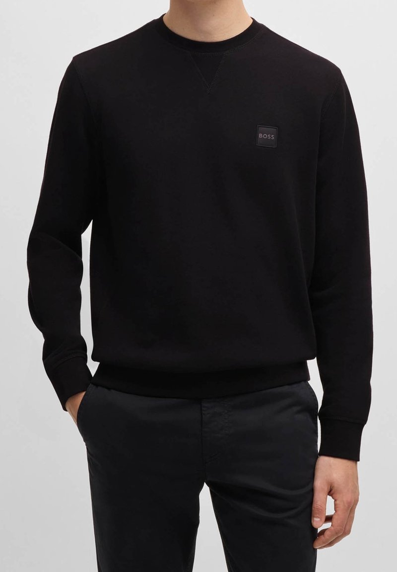 Homme portant un sweat-shirt noir à manches longues avec un petit logo BOSS sur la poitrine et un pantalon foncé, mains dans les poches, debout devant un fond uni.