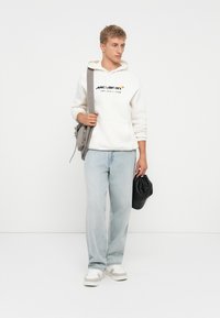 Ung mand iført en hvid McLaren hoodie, lyseblå jeans, hvide sneakers, bærende en grå skuldertaske og holdende en sort kasket.