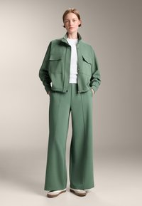 Giacca verde con due tasche frontali, abbinata a pantaloni a gamba larga e una maglia bianca aderente. Texture liscia, silhouette moderna, design casual.