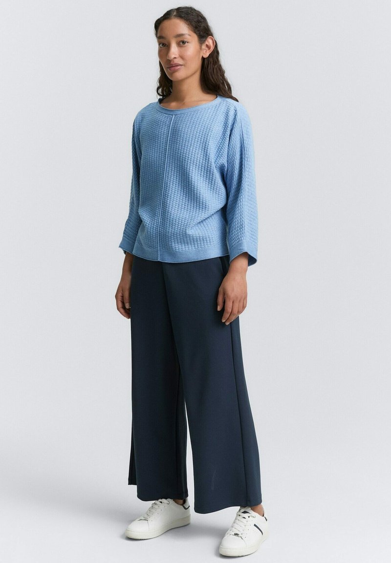 Jasnoniebieski sweter o fakturze z okrągłym dekoltem i rękawami 3/4, w połączeniu z szerokimi granatowymi spodniami i białymi sneakersami.