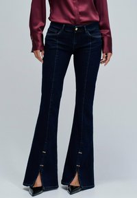 Mujer con jeans oscuros de campana con aberturas delanteras y broches dorados, combinados con tacones negros puntiagudos y una blusa de satén burdeos.