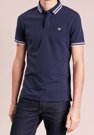 Mężczyzna w granatowej koszulce polo z krótkim rękawem, białym paskiem na kołnierzyku i mankietach rękawów, w połączeniu z ciemnoniebieskimi dżinsami.