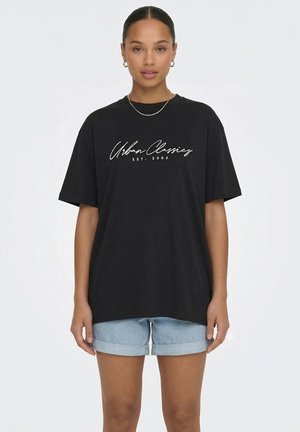 Kvinde iført sort oversized T-shirt med hvidt "Urban Classics EST. 2005" tekst og lyseblå opsmøgede denimshorts, stående forfra.