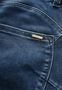 Tummansiniset farkut, joissa on sileä pinta, kontrastiväriset ompeleet ja pieni merkkilappu taskussa.