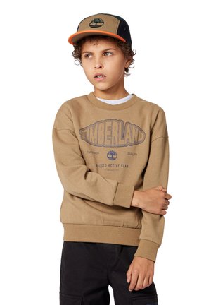 Sudadera marrón con el logo de "Timberland" en azul, cuello redondo y puños de canalé. Llevada con una gorra negra y marrón que presenta un detalle del logo.