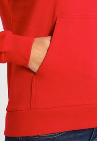 Czerwona bluza z kangurzą kieszenią i ściągaczami przy mankietach. Materiał wydaje się gładki, ma miękką teksturę i dopasowany krój w talii.