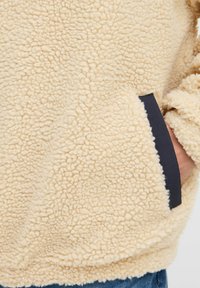 Beige fleece jacket with a soft, textured surface, featuring black trimmed pockets and a relaxed fit. Ideal for warmth and comfort.