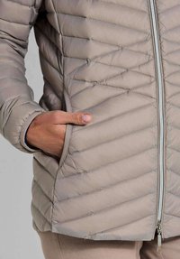 Persoon in een licht taupe gewatteerde jas met een hand gedeeltelijk in de zijzak nabij de voorkant met rits.