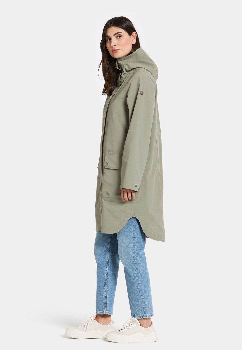 Hellgrüne Parka mit Kapuze, Fronttaschen und geschwunghem Saum. Kombiniert mit blauen Jeans und weißen Turnschuhen. Hergestellt aus einem glatten, wasserabweisenden Material.