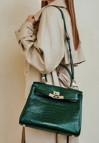 Zielona torebka z tłoczonym wzorem krokodyla o wyraźnej strukturze, ze złotymi akcentami i odpinanym paskiem, oparta o beżowy płaszcz.
