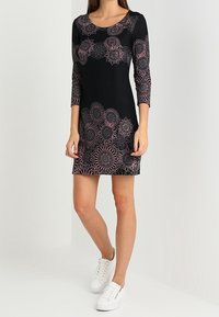 Svart klänning med ett blommönster i spets som är rosa. Den har en figursydd design med trekvartsärmar och hög rund halsringning, kombinerad med vita sneakers.