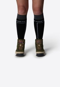 Botas de senderismo verdes y color arena con cordones morados y amarillos, combinadas con calcetas negras hasta la rodilla con acentos blancos y un logo.