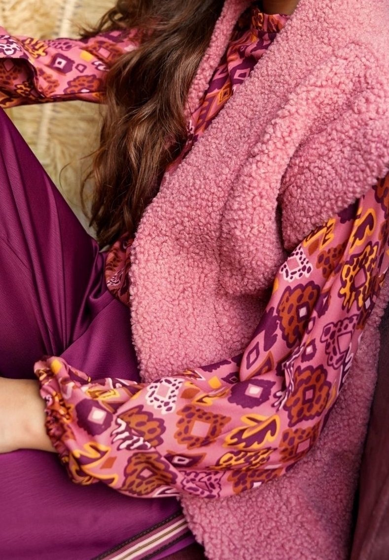 Veste en polaire rose pâle associée à un chemisier à manches longues à motifs floraux violets, orange et jaunes.