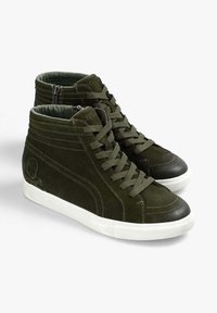Zapatillas altas de ante de color verde oscuro con suelas blancas, que cuentan con cordones, costuras decorativas y un pequeño emblema en el lateral.