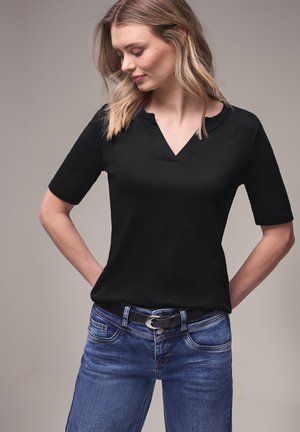 Kobieta o blond włosach, ubrana w czarną koszulkę z krótkim rękawem i niebieskie dżinsy z czarnym paskiem, patrząca w dół, z rękami za plecami.