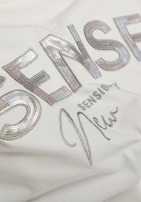 Silberne Pailletten- und Fadenstickerei auf weißem Stoff mit der Aufschrift "DENSEL", darunter kleinerer Text "SENSIBLY" und eine kursiv geschriebene Unterschrift.