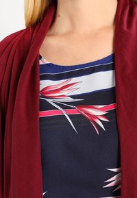Bordowy kardigan na granatowym topie z kwiatowym wzorem. Top ma poziome paski w odcieniach niebieskiego i różowego, a także białe i różowe akcenty w postaci kwiatów.