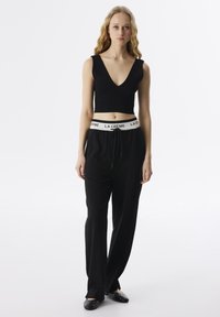 Zwarte geribbelde crop top met v-hals, gecombineerd met een losse zwarte broek met een witte "LA CRÈME" logo tailleband. Platte zwarte schoenen.