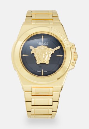 Gold metal watch with a black face featuring a lion head detail, hour markings, and "Versace" logo; secured with a bracelet link band.